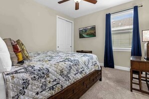 Ein Schlafzimmer mit Bett, Deckenventilator, Fenster mit Vorhängen, Nachttisch und einem an der Wand hängenden Bild.