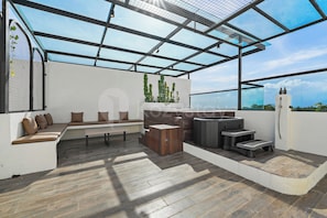 A rooftop terrace with a glass ceiling, a seating area with cushions, a wooden table, and a hot tub.
