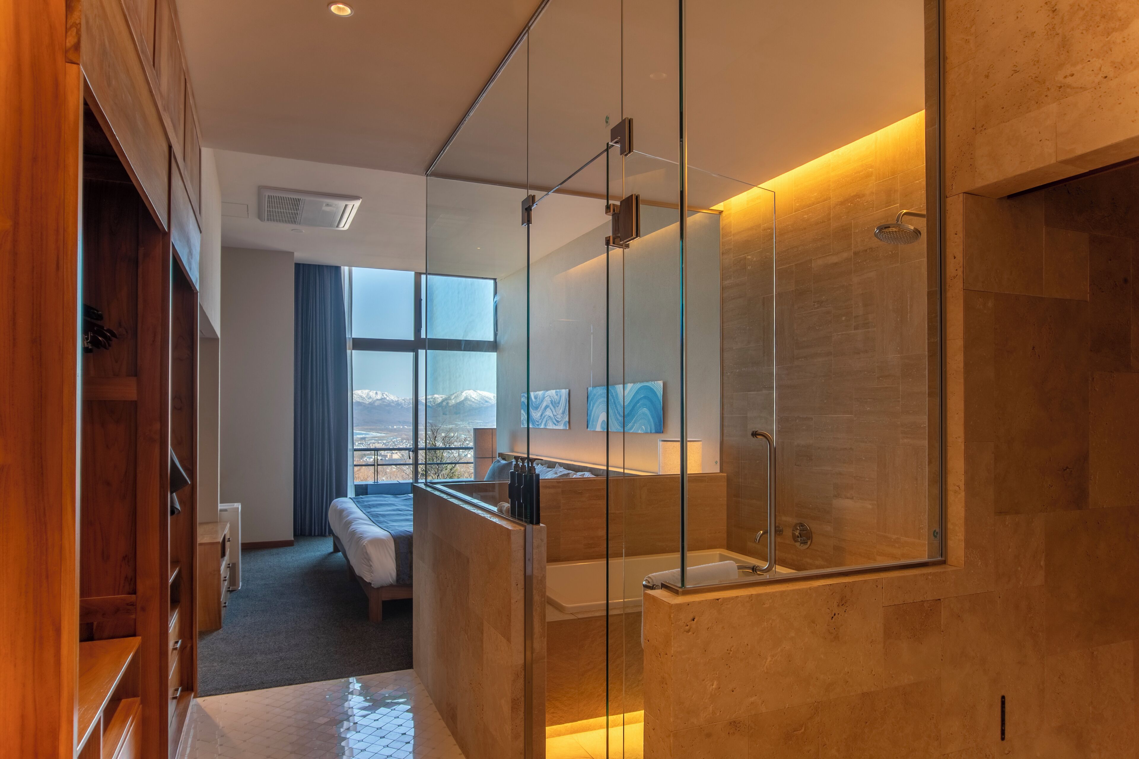 Una habitación de hotel moderna con cabina de ducha de vidrio, una cama, vistas a montañas y un armario de madera.