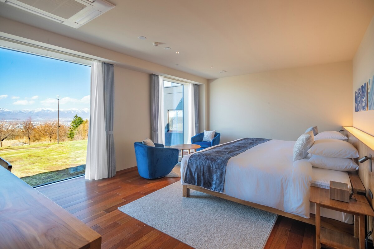 A modern hotel room with a large bed, a seating area with a blue chair, a small table, and a view of a mountainous landscape through large windows.