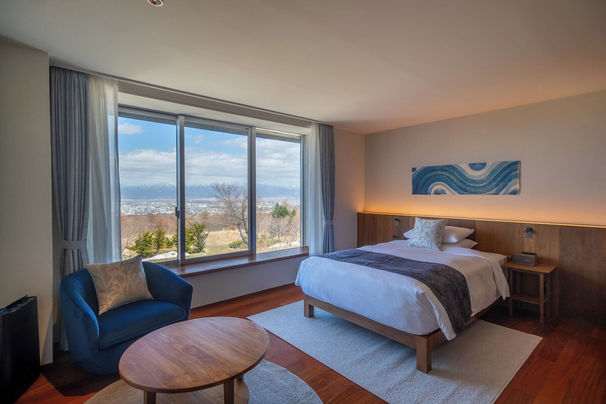 A hotel room with a large bed, a bedside table with a lamp, a chair, a small round table, and a view of the mountains through the window.