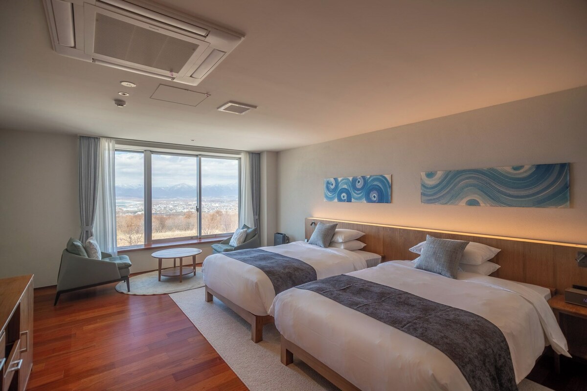 A hotel room with two beds, a seating area with a table, and a large window offering a mountain view.