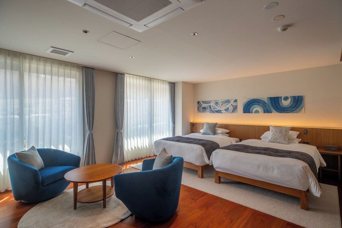 A hotel room with two beds, a sofa, a small table, and a large window with curtains.