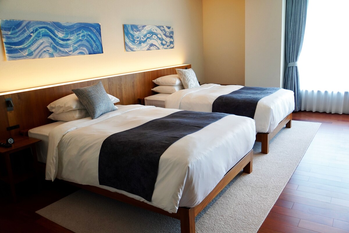 A hotel room with two beds, a wooden headboard, a nightstand, a lamp, and two abstract paintings on the wall.