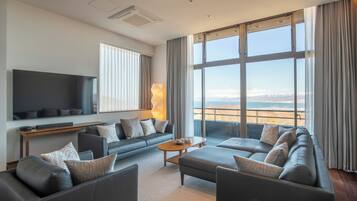 A modern living room with a large flat-screen TV, a sofa set, a coffee table, and a view of the sea.