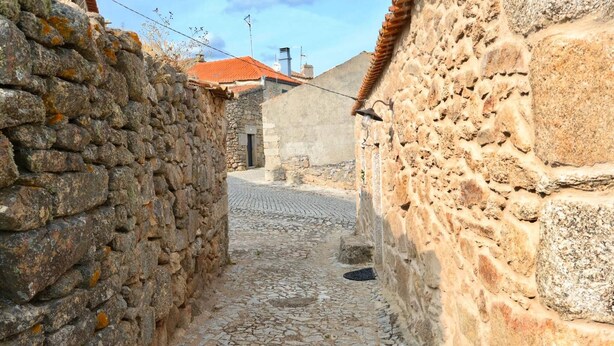 Uma rua de paralelepípedos estreita, ladeada por muros de pedra, que conduz a um edifício com telhado de telhas vermelhas.