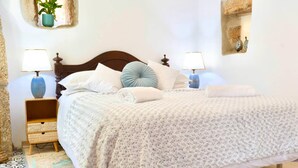 Uma cama bem arrumada com colcha estampada, duas luminárias de cabeceira, uma mesa de cabeceira com cesto de fibra e um vaso de planta na parede.