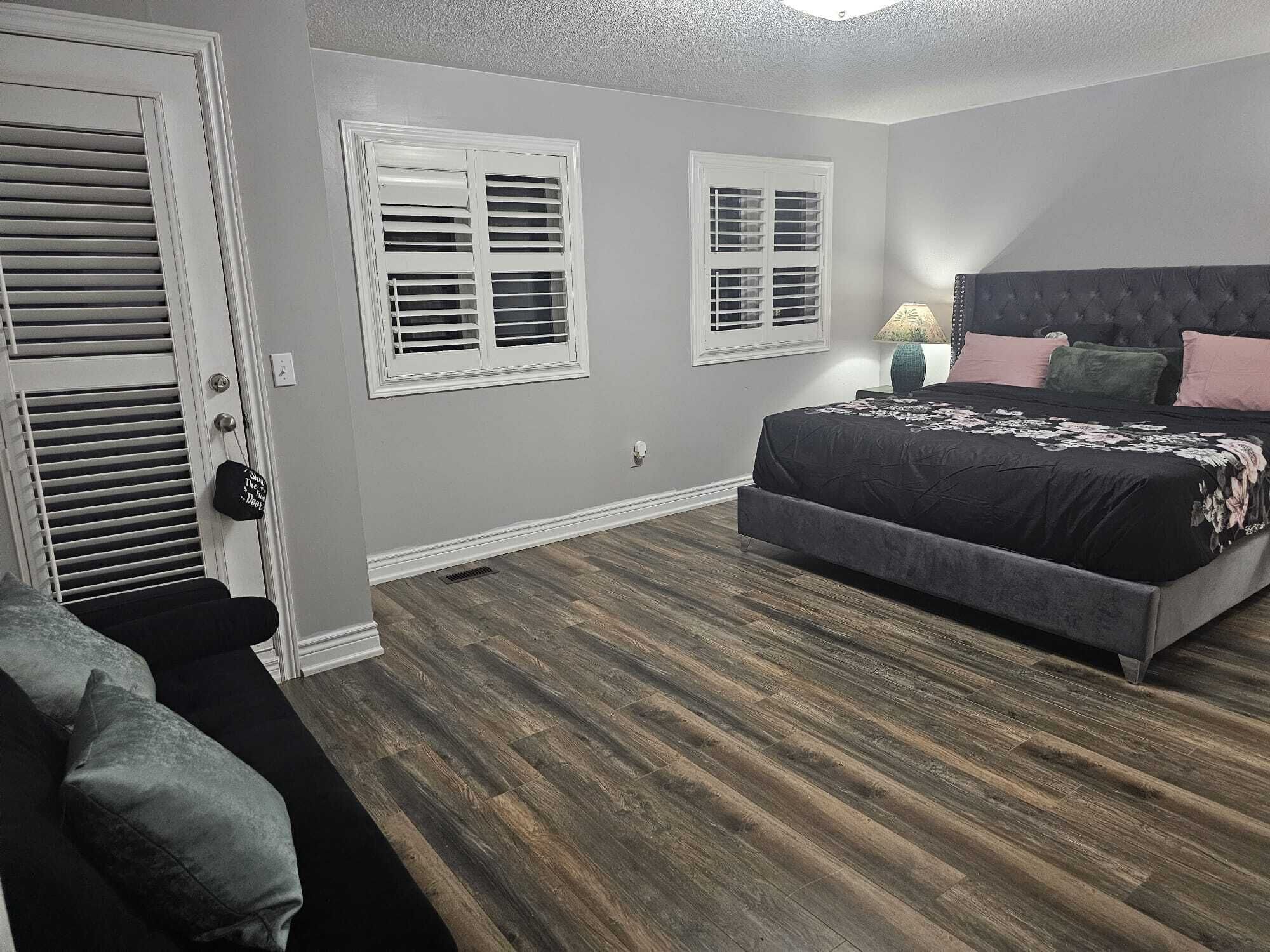 Une chambre à coucher avec un lit, deux fenêtres, une porte et une table de chevet avec une lampe.
