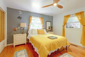 Une chambre à coucher comprenant un lit, une table de chevet, une lampe, une fenêtre avec des rideaux et un ventilateur de plafond.