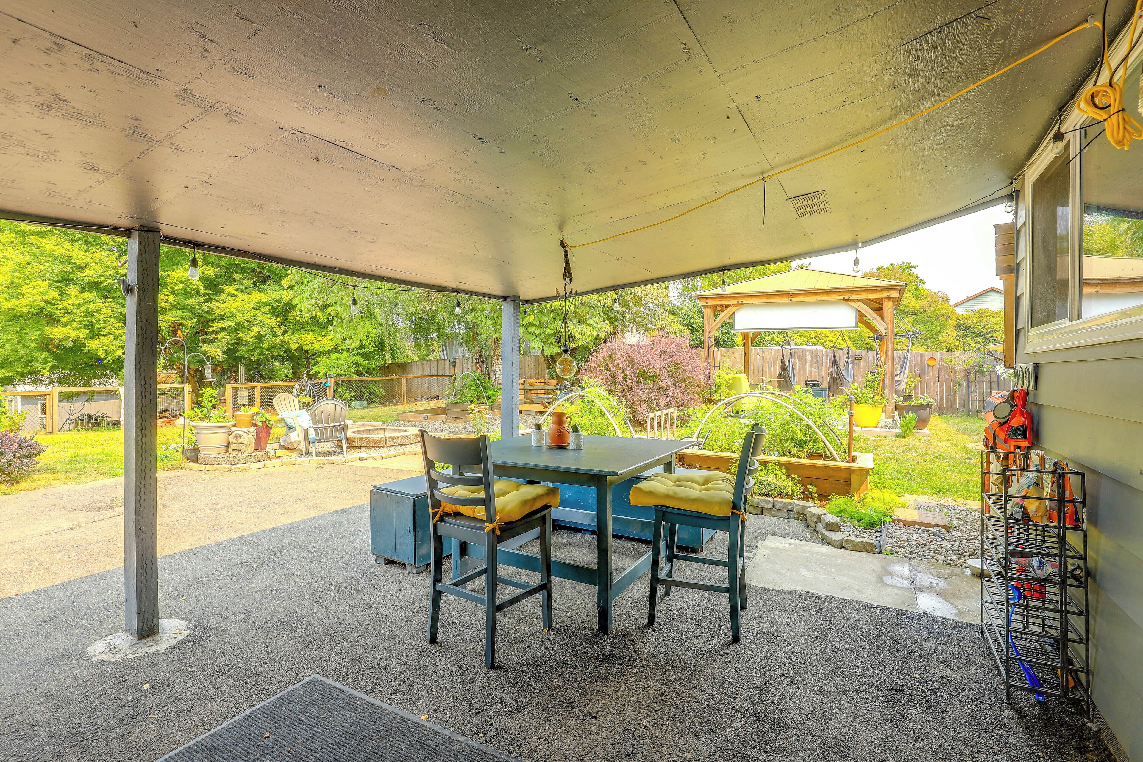 A covered patio area with a table and chairs, a garden view, and a metal rack with bags.