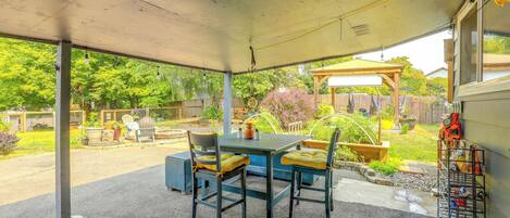 A covered patio area with a table and chairs, a garden view, and a metal rack with bags.