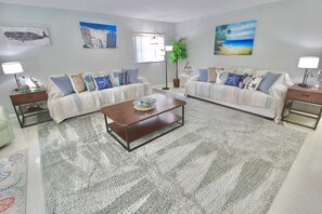 A living room with two sofas, a coffee table, and a rug. There are paintings on the walls and a plant in the corner. - The Seaside Serenity at Thumb Point - Hutchinson Island - Prime Location - Pool Home! (Fort Pierce)