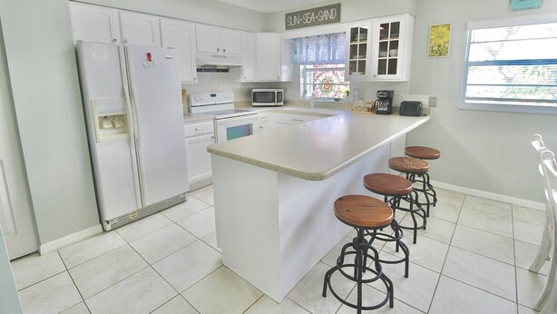 A kitchen with a central island, bar stools, a refrigerator, a microwave, and a coffee maker. - The Seaside Serenity at Thumb Point - Hutchinson Island - Prime Location - Pool Home! (Fort Pierce)