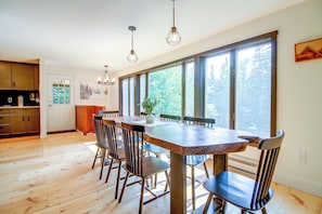 A dining area with a wooden table and chairs, large windows with a view of trees, and a kitchen with wooden cabinets. - 1 Mi to Mount Snow! Classic Chalet w/ Hot Tub (West Dover)