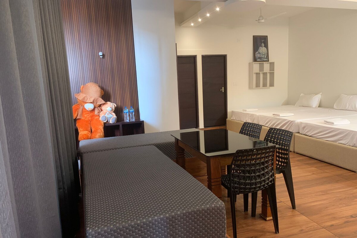 Habitación de hotel moderna con cama, una mesa de comedor con sillas, un banco y un gran peluche de oso decorativo.