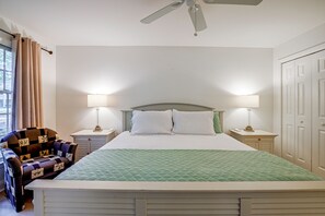 Un dormitorio con una cama, dos mesitas de noche, una silla, un ventilador de techo y una ventana con cortinas.