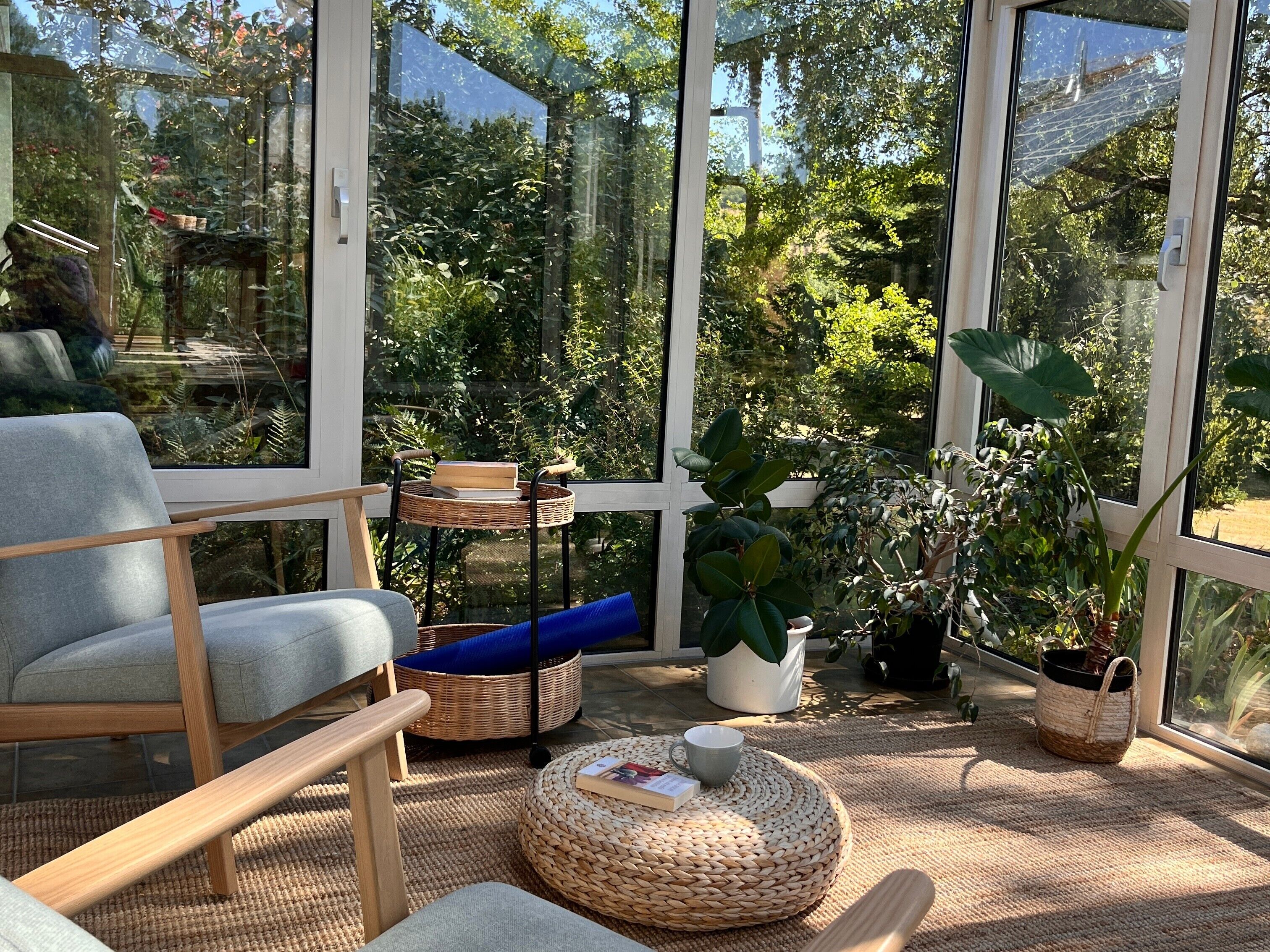 Une pièce moderne avec de grandes fenêtres, un canapé gris, des meubles en osier et des plantes en pot.
