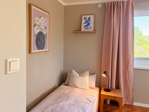 A bedroom with a bed, pillows, a nightstand, a lamp, a window with curtains, and two framed artworks on the wall.