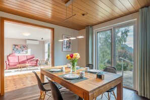 A dining area with a wooden table set for four, a pink sofa, and a large window with a view of trees.