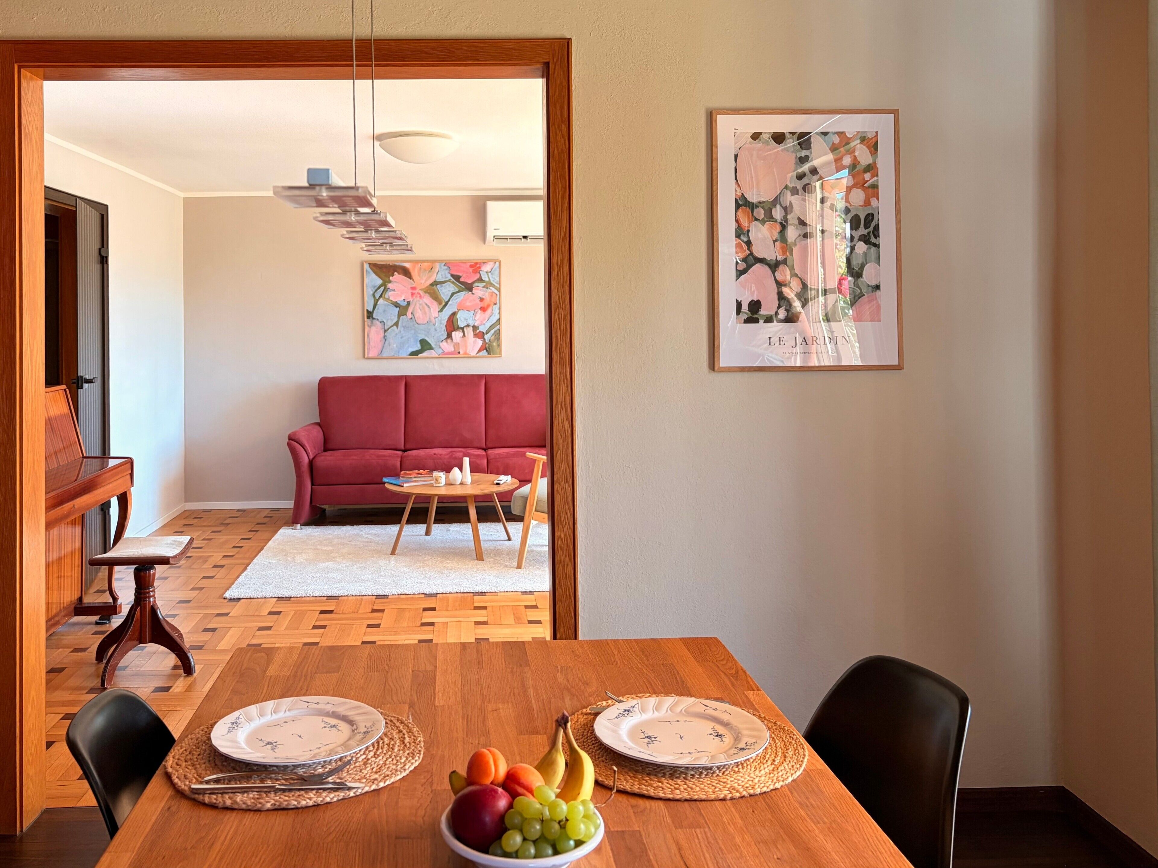 Un coin repas avec une table en bois dressée pour deux personnes, un panier de fruits et, au fond, un canapé rouge.