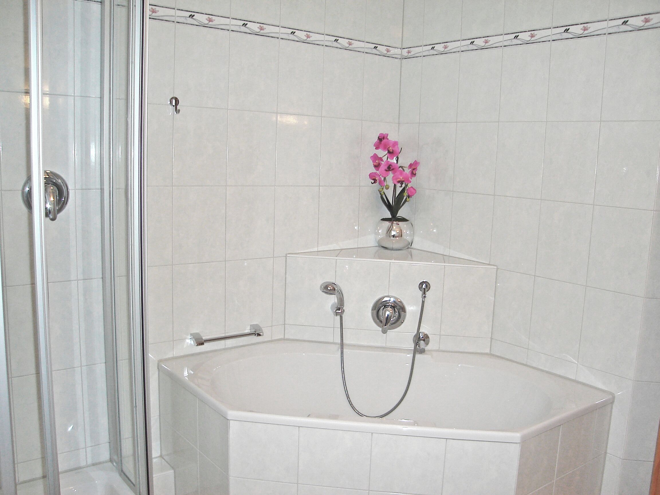 A bathroom with a glass shower enclosure, a bathtub with a showerhead, and a vase with pink flowers on a shelf.