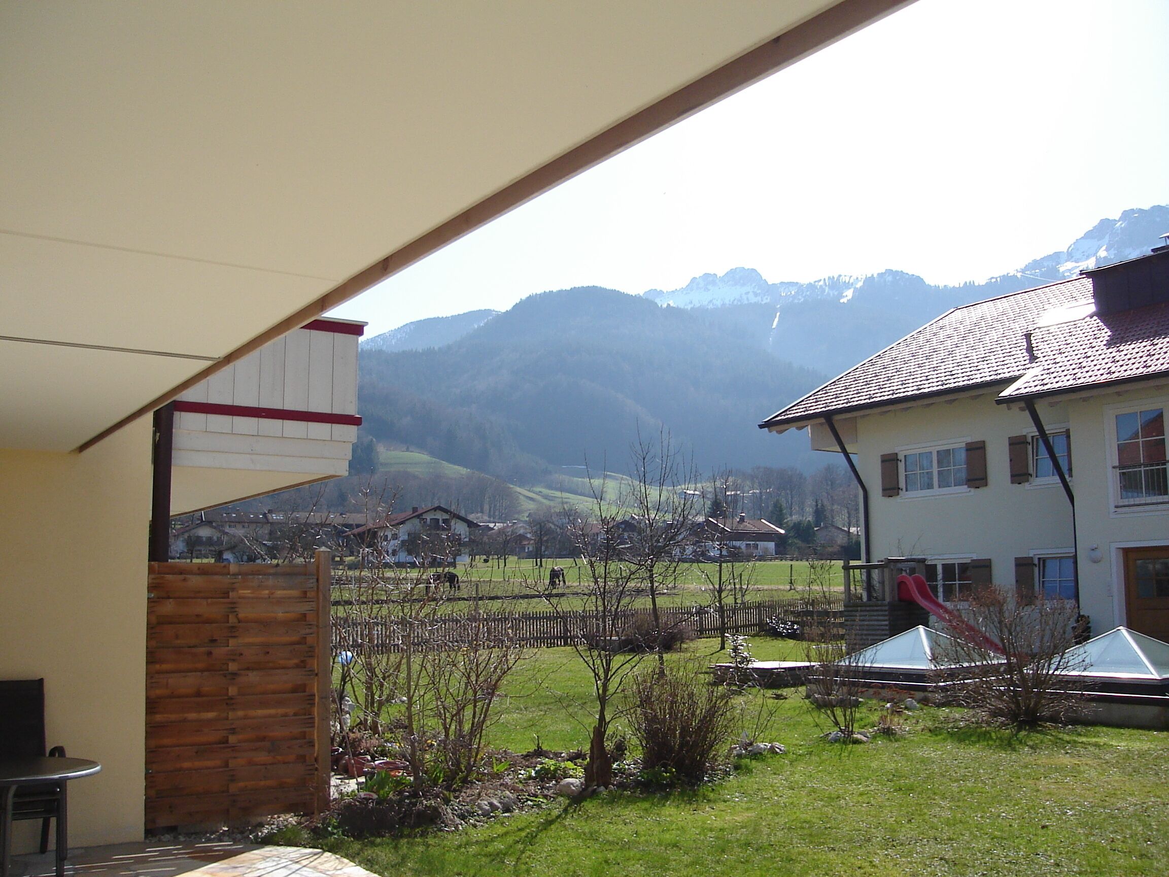 A covered outdoor area with a view of a mountain range, a wooden fence, and a building with a tiled roof.