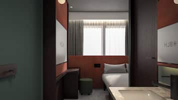 Habitación de hotel compacta con una cama, un tocador con espejo, un escritorio pequeño y una ventana con cortinas.