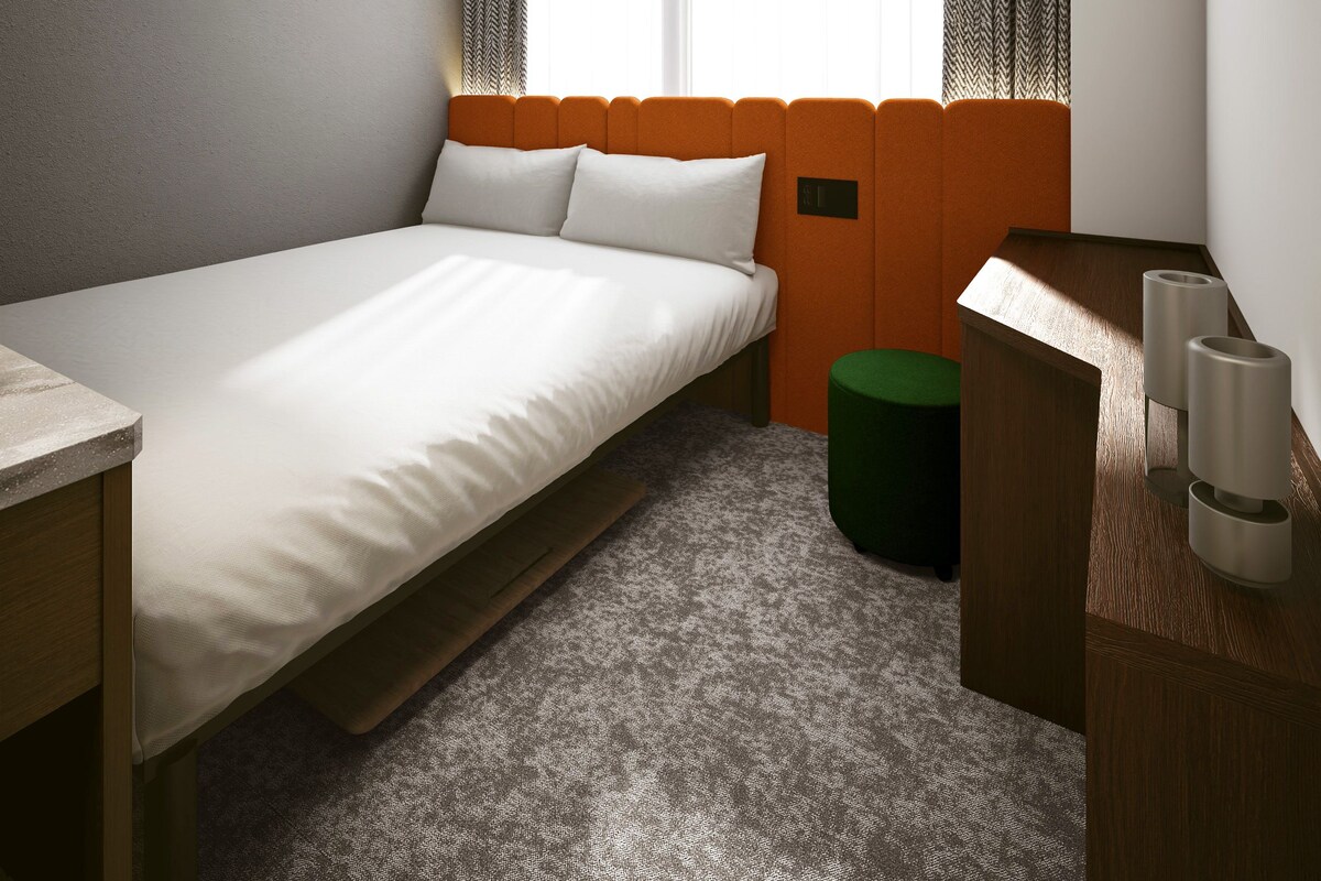 Una habitación de hotel moderna con una cama individual, un pequeño banquillo verde, una mesita de noche de madera y una ventana con cortinas estampadas.