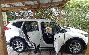 A white SUV parked under a wooden structure with a metal roof, surrounded by greenery. - Beautiful 3 bedroom apartment with pool on Av (Santo Domingo)