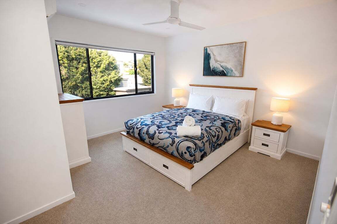 A bedroom with a large bed, two bedside tables, a ceiling fan, a window with a view of trees, and a painting on the wall.