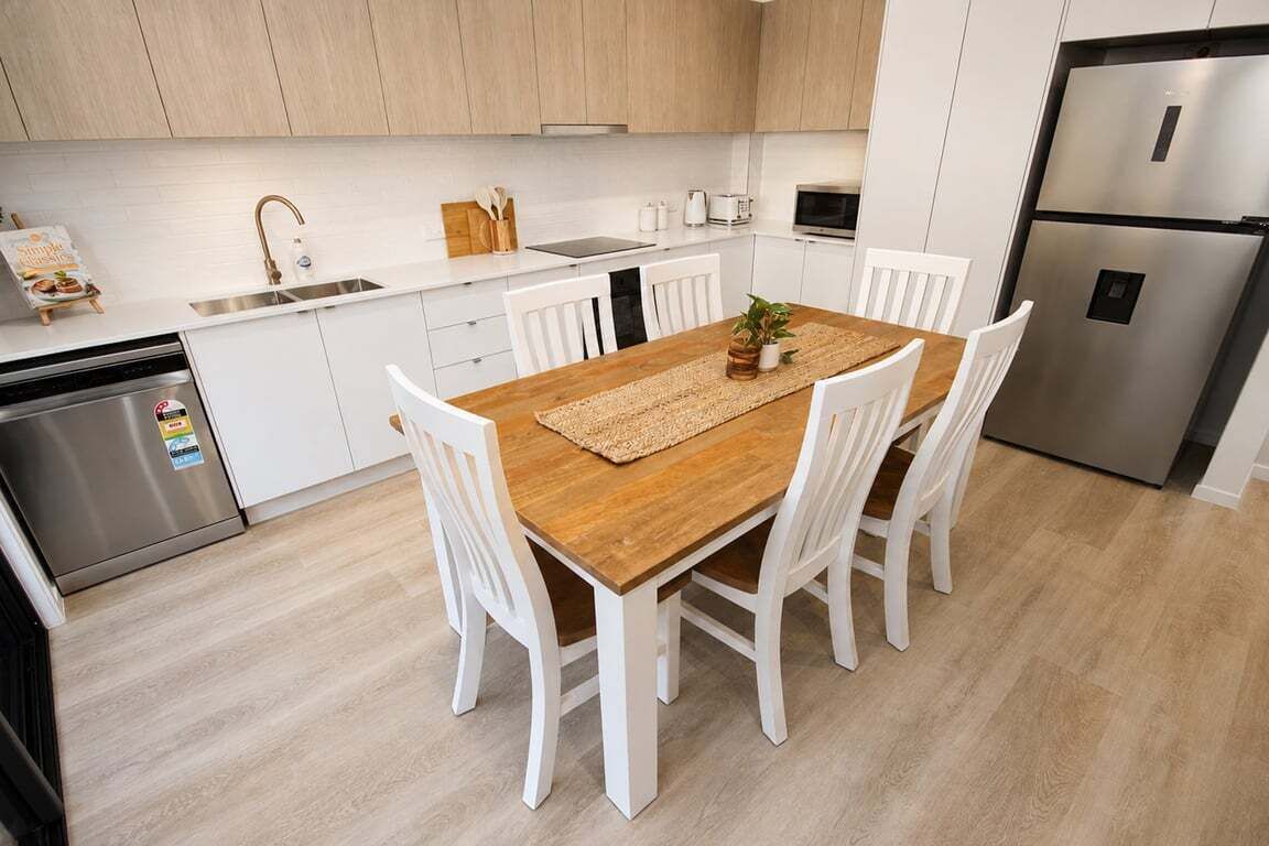 A modern kitchen with white cabinetry, a wooden dining table, and a stainless steel refrigerator.
