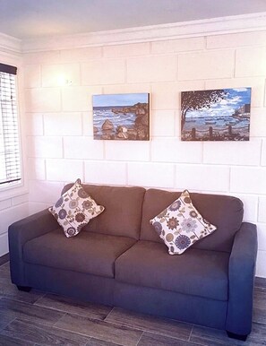 A dark-colored sofa with floral-patterned cushions against a white brick wall. Two framed paintings depicting coastal scenes are hung above the sofa. - Panthera Terra #1 - By ZenBreak (Mount Standfast)