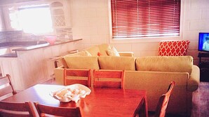 Ein Wohnzimmer mit einer Sofagarnitur, einem Esstisch mit Stühlen, einem Fernseher und einer Küchenzeile mit einem Fenster.