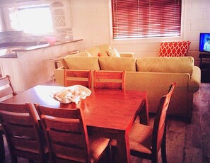 Ein Wohnzimmer mit einer Sofagarnitur, einem Esstisch mit Stühlen, einem Fernseher und einer Küchenzeile mit einem Fenster.