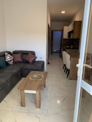 A modern living room with a sectional sofa, a small wooden coffee table, and a kitchen area visible through an open door. - La prestigia Hurghada Apartment (Red Sea Governorate)