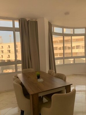 A room with a wooden table and chairs, large windows with curtains, and a view of buildings outside. - La prestigia Hurghada Apartment (Red Sea Governorate)