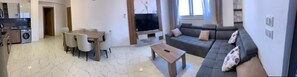 A modern living room with a sofa, a coffee table, and a TV. There is a dining area with chairs and a table. The room has a tiled floor. - La prestigia Hurghada Apartment (Red Sea Governorate)