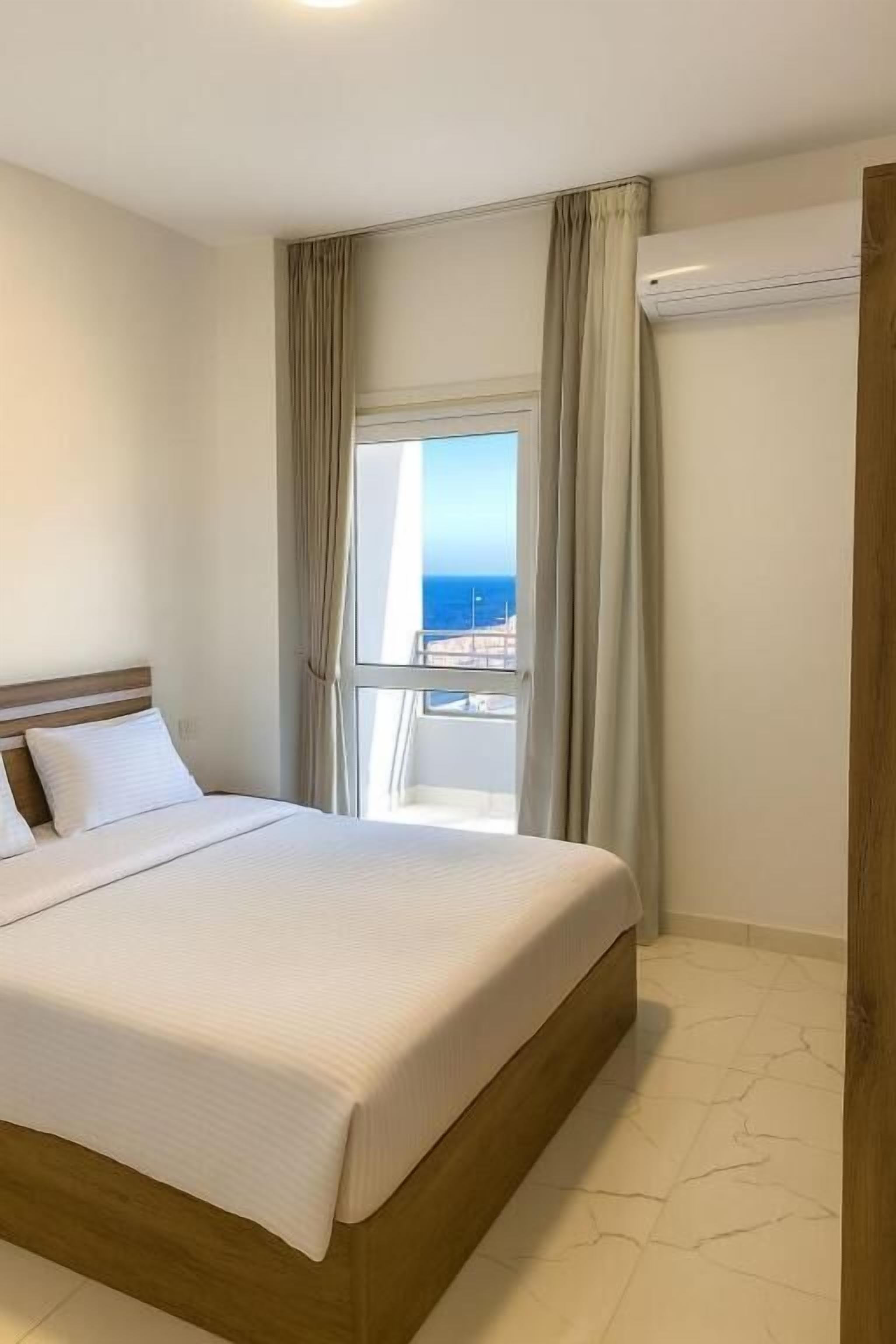 Quarto de hotel com uma cama grande, janela com cortinas e vista para o mar.