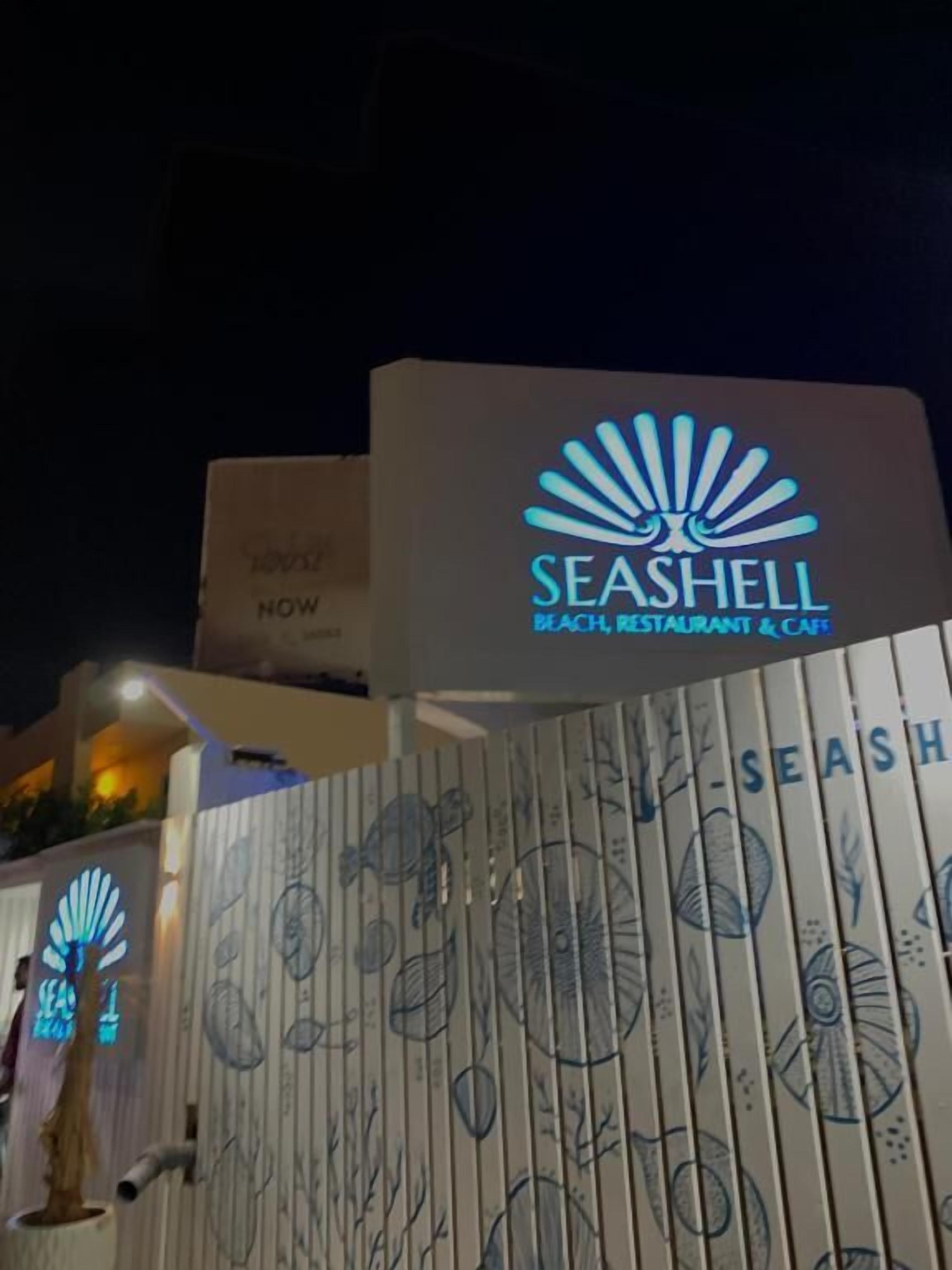 Opastekyltti, jossa lukee ”SEASHELL BEACH RESTAURANT & CAFE”, ja jossa on logon sekä koriste-elementtejä.