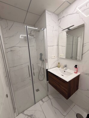 Un baño moderno con ducha envuelta en vidrio, un lavamanos blanco con tocador de madera y pared con estampado de mármol.