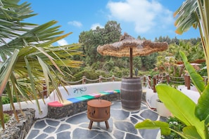 Un patio con suelo de piedra, un barril de madera, un parasol de paja y un área de estar colorida.