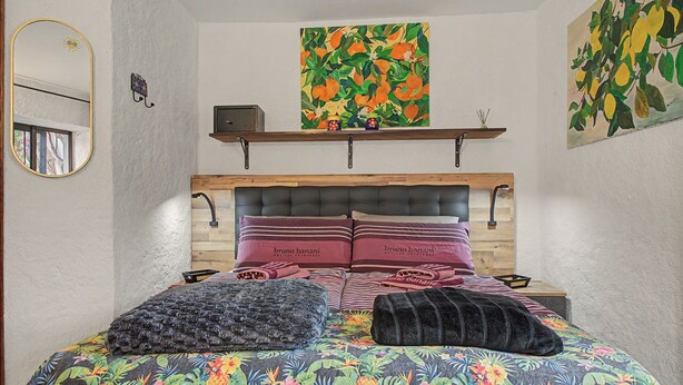 A bedroom with a bed, pillows, a headboard, a shelf with decorative items, and a painting on the wall.