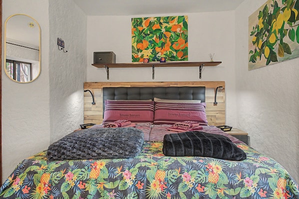 Un dormitorio con cama, almohadas, cabecera, estante con objetos decorativos y un cuadro en la pared.