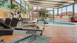 A sunlit indoor swimming pool area with exercise equipment, lounge chairs, and potted plants.