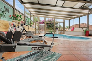 Un área de piscina interior iluminada por el sol, con equipo de ejercicio, sillones de descanso y plantas en macetas.