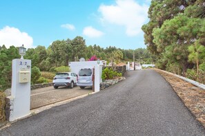 Un camino pavimentado que conduce a una entrada con puerta y dos coches estacionados, rodeado de follaje y árboles.