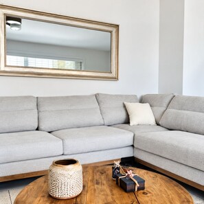 A living room with a grey sectional sofa, a round wooden coffee table, a woven basket, and a framed mirror on the wall. - Sea la  Vie (Saronida)