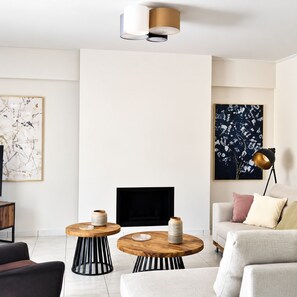 A modern living room with a fireplace, two round wooden tables with vases, a sofa with cushions, and artwork on the walls. - Sea la  Vie (Saronida)