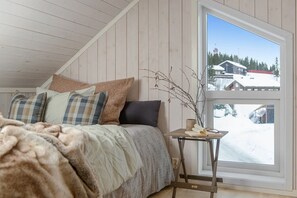 Ein Schlafzimmer mit einem Bett, Kissen, einem Nachttisch mit Buch und Brille sowie einem Fenster mit Blick auf Schnee.