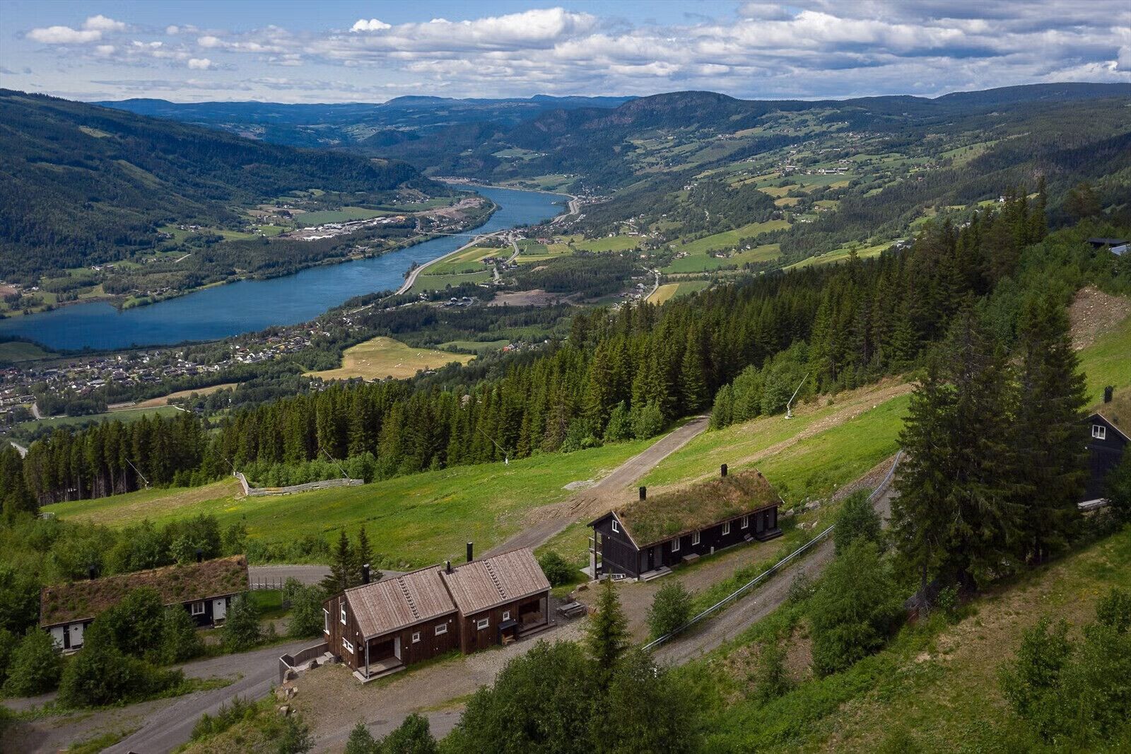 Ein abgelegenes Häuschen, verbunden durch einen Feldweg, umgeben von dichtem Wald und einem See in der Ferne.